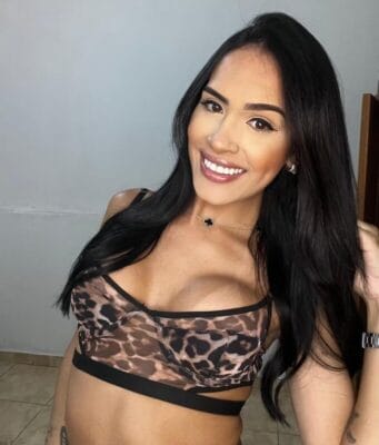 Smiling woman with long black hair wearing a leopard-print bralette, taking a selfie indoors.