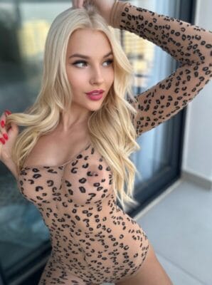 Blonde woman wearing a leopard-print sheer bodysuit posing with one arm raised near a window.