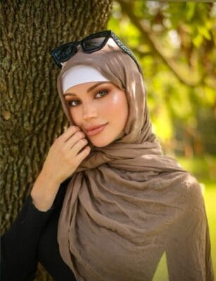 Woman wearing a beige hijab with a white inner headband, leaning against a tree in a sunny park, sunglasses resting on her head.