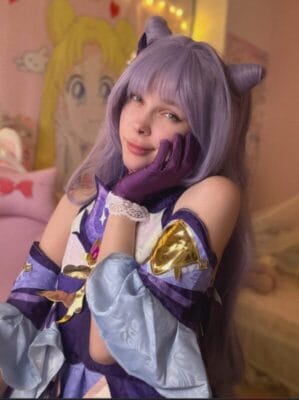 Cosplayer with lavender wig and horned headband posing with chin resting on gloved hand, wearing a purple and light blue fantasy costume in a pink, anime-themed bedroom.