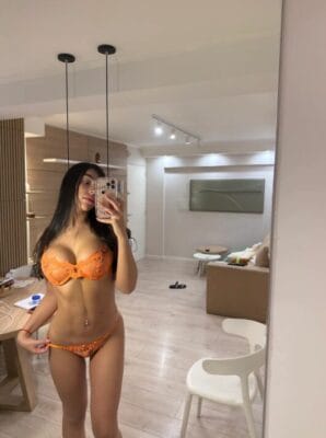 Woman in orange lace lingerie taking a mirror selfie in a modern living room with a beige sofa and minimalist decor.