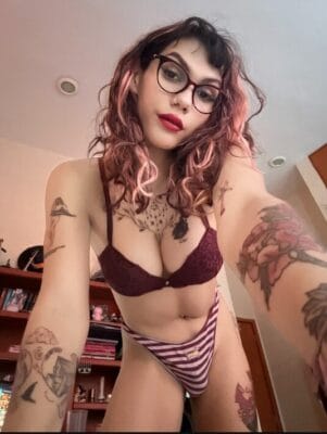 Self-portrait of a tattooed person wearing burgundy lingerie and striped underwear, with pink-dyed hair, glasses, and red lipstick taking a selfie in a bedroom.