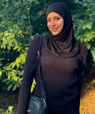 Smiling woman wearing a black hijab and brown ribbed sweater, standing outdoors with green foliage and a crossbody bag.