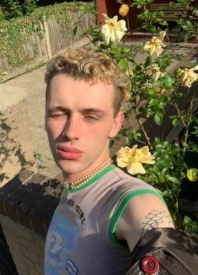 Young man taking a selfie outside beside a rose bush, wearing a gray sleeveless shirt with green trim, a beaded necklace, nose ring, and a shoulder tattoo.