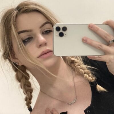Young woman with blonde braided pigtails taking a mirror selfie with a white iPhone, wearing a necklace and minimal makeup.