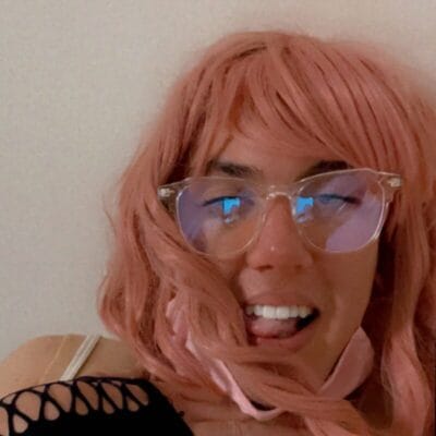 Person with a pink wig and clear-framed glasses winking and sticking out their tongue playfully against a light background.