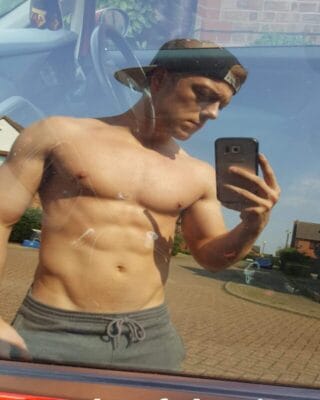 Shirtless man wearing a backwards cap takes a mirror selfie in a car with a smartphone.