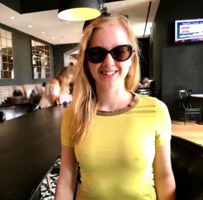 Smiling woman with long blond hair wearing black sunglasses and a yellow shirt in a cafe setting.