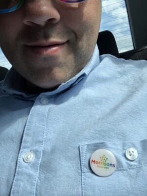 Close-up of a person wearing a light blue shirt with a Morisons badge on the chest pocket, seated in a car.