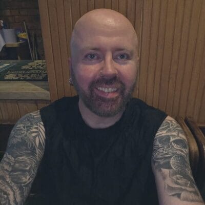 Smiling bald man with a beard and nose ring, wearing a black sleeveless shirt, tattoos on both arms in a wood-paneled room.