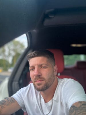 Man with short hair and beard wearing a white t‑shirt sits in a car, taking a selfie; red car seats and window visible.