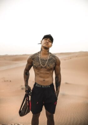 Shirtless man with numerous tattoos standing in a desert, holding a bag and wearing a cap and chains.