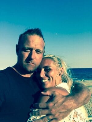 A couple hugs at the beach, the man with a dark shirt and the woman smiling as waves roll in the background on a sunny day.